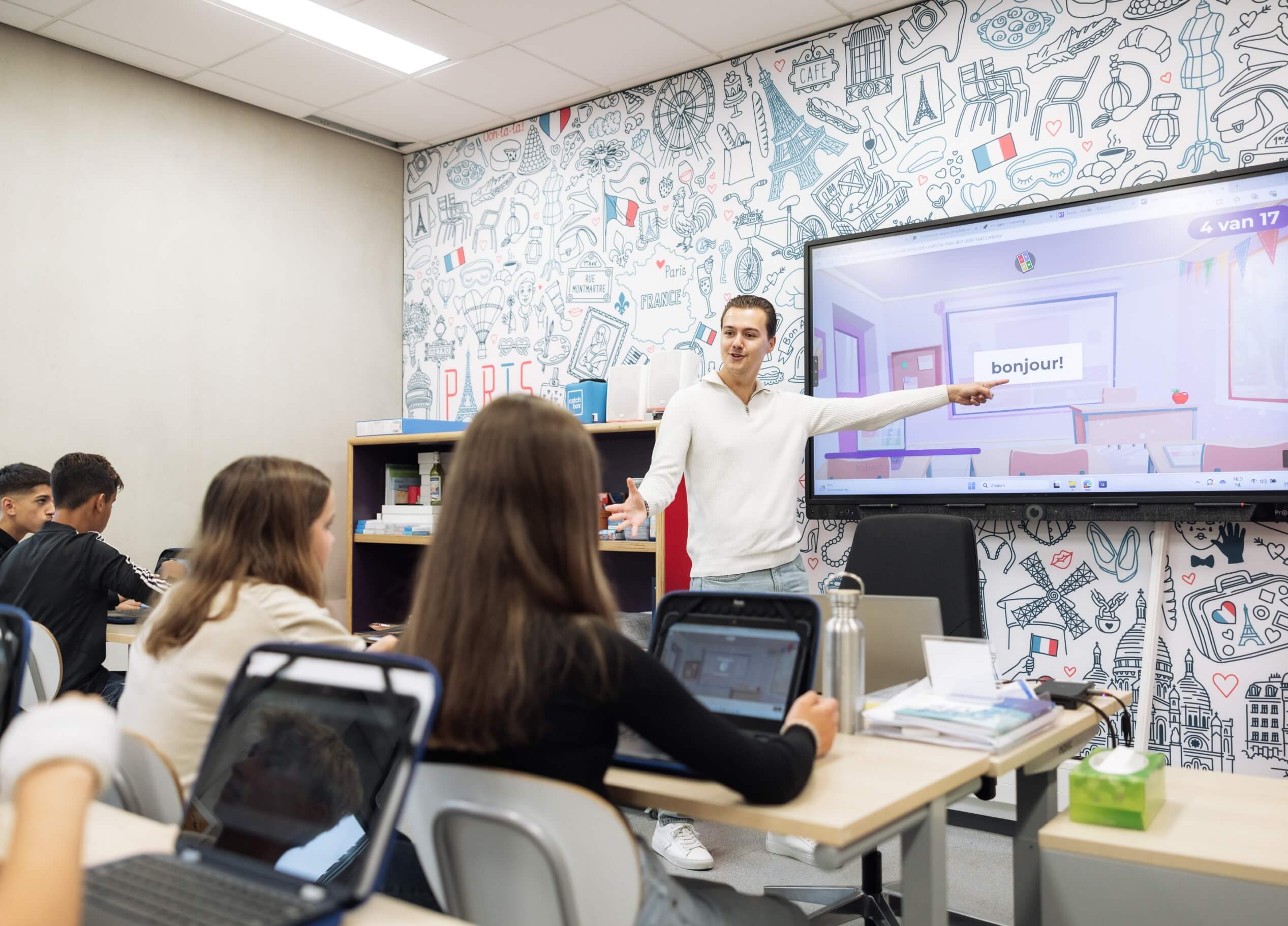 Een leraar wijst naar een "bonjour" op het scherm, terwijl leerlingen op laptops werken.