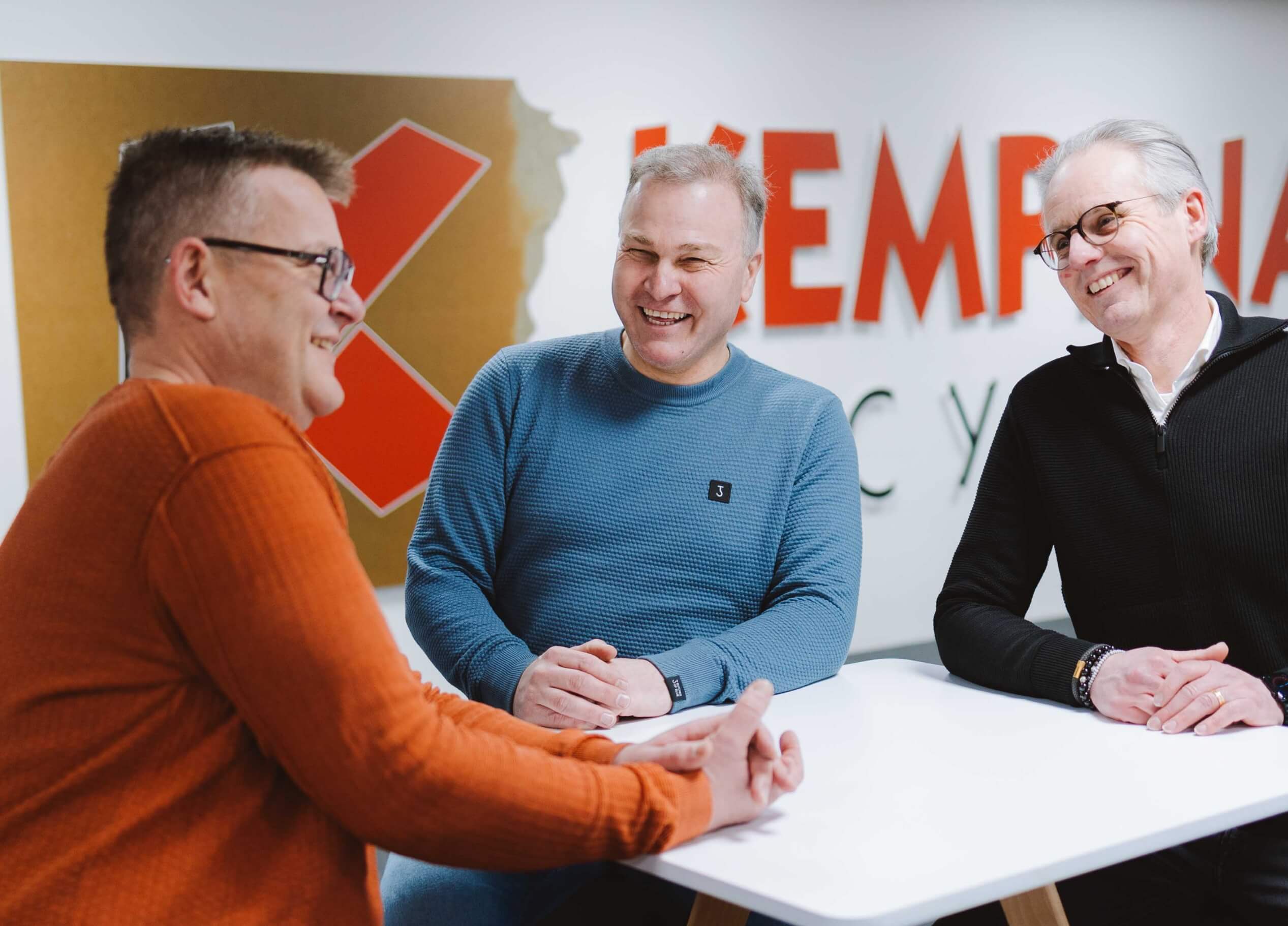 Drie mannen kletsen en lachen rond een witte tafel in een strak, modern kantoor.
