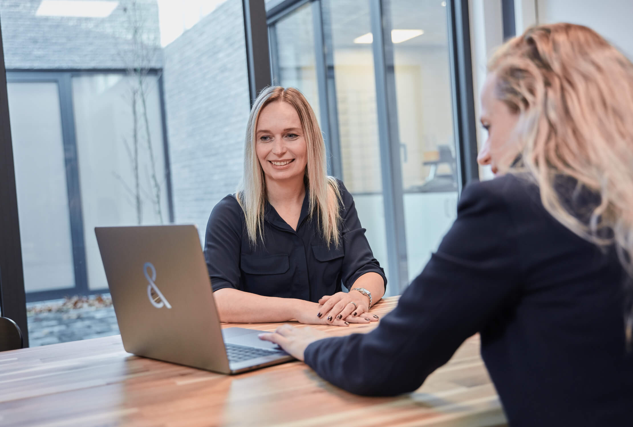 Twee aan een houten tafel met laptops: één lacht, één geconcentreerd, kantoorsfeer.