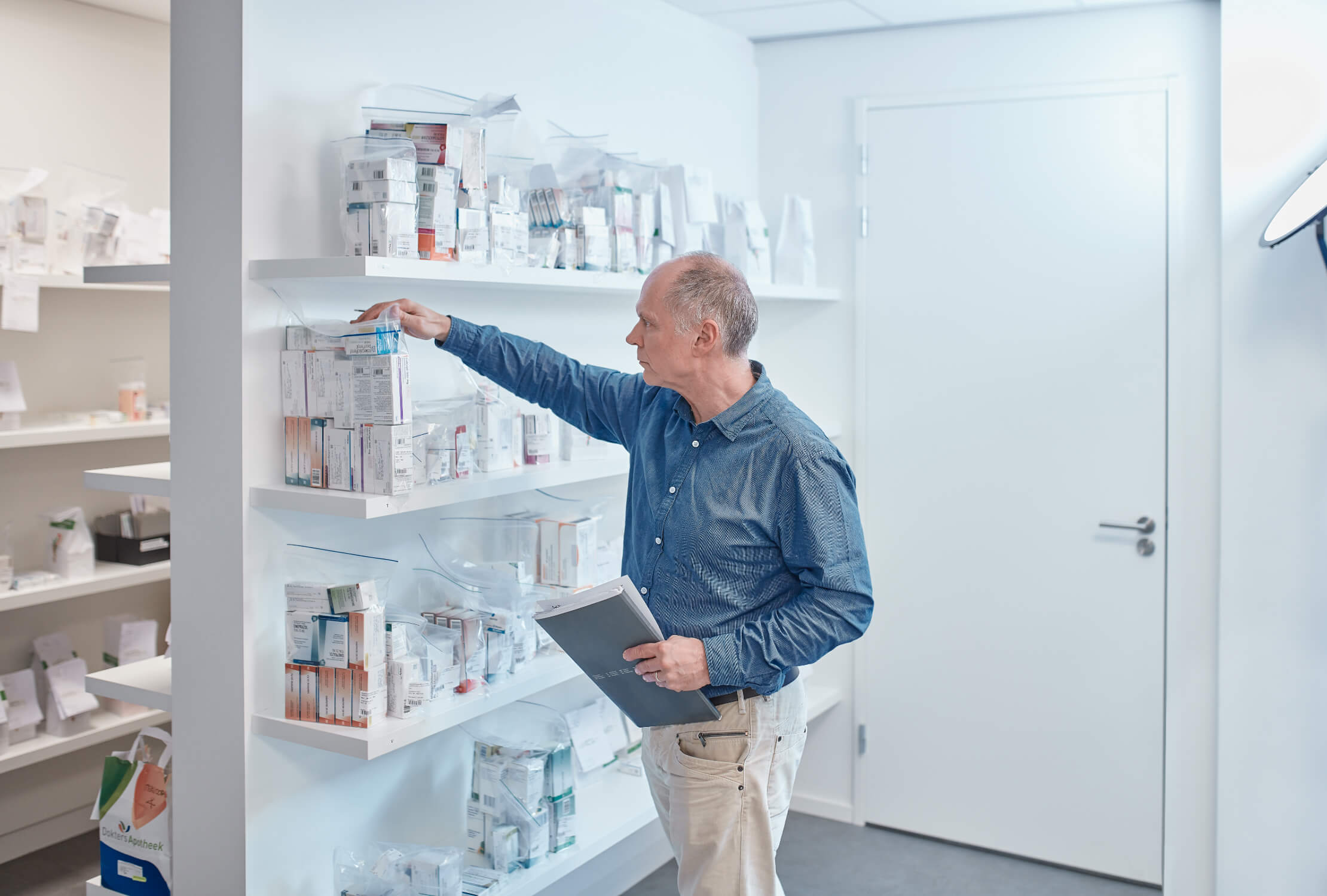 Man in blauw shirt sorteert medische benodigdheden op planken, klembord in de hand, in een lichte kamer.