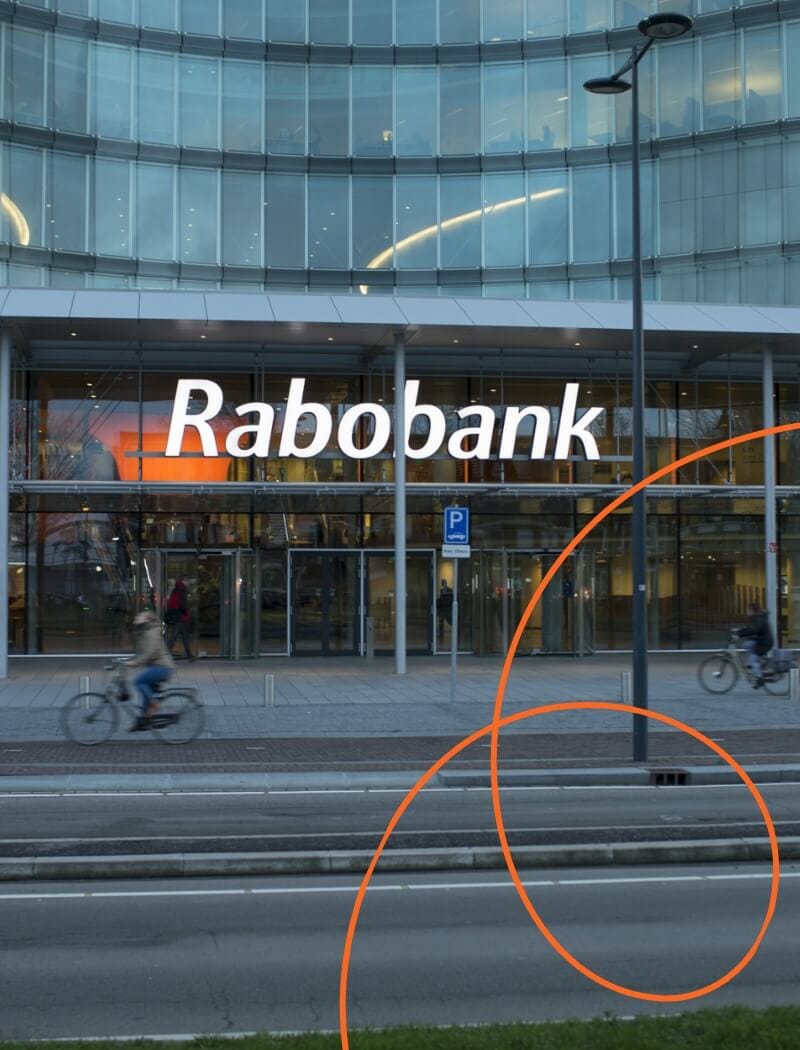 Glazen Rabobankgebouw, fietsers rijden voorbij; gebogen oranje lijnen op de voorgrond.