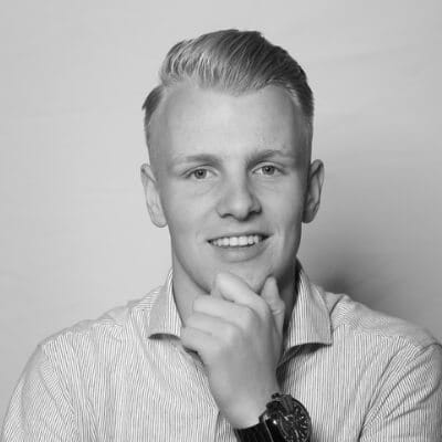 Een jongeman met kort blond haar lacht, kin op de hand, in een gestreept shirt en horloge. Zwart-witfoto.