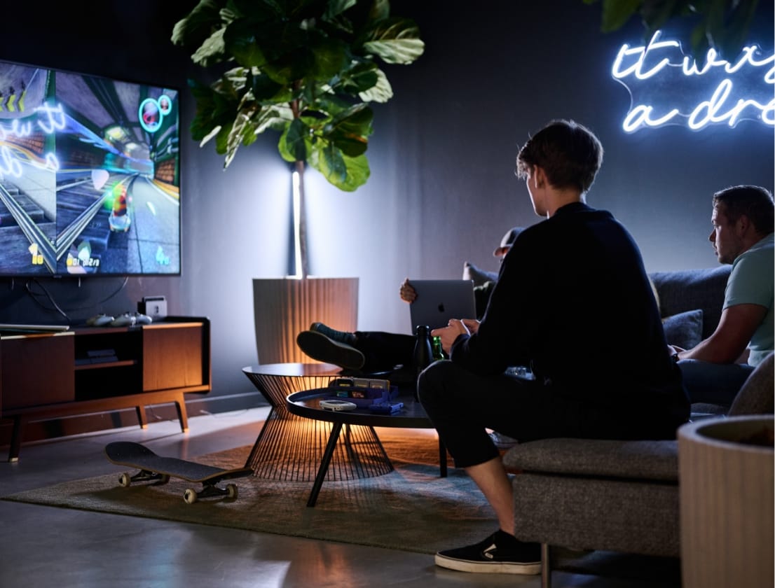 Gamers in een gezellige woonkamer met een grote tv, een skateboard en een neonreclame.