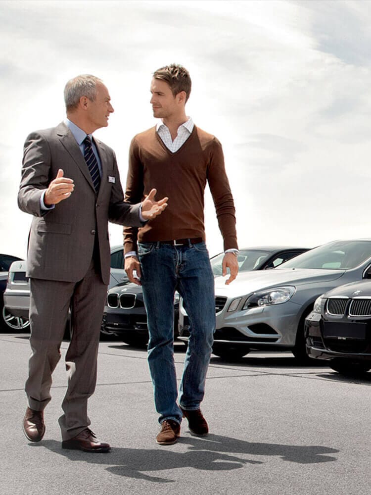 Twee mannen kletsen en lopen synchroon langs geparkeerde auto's onder een bewolkte lucht.