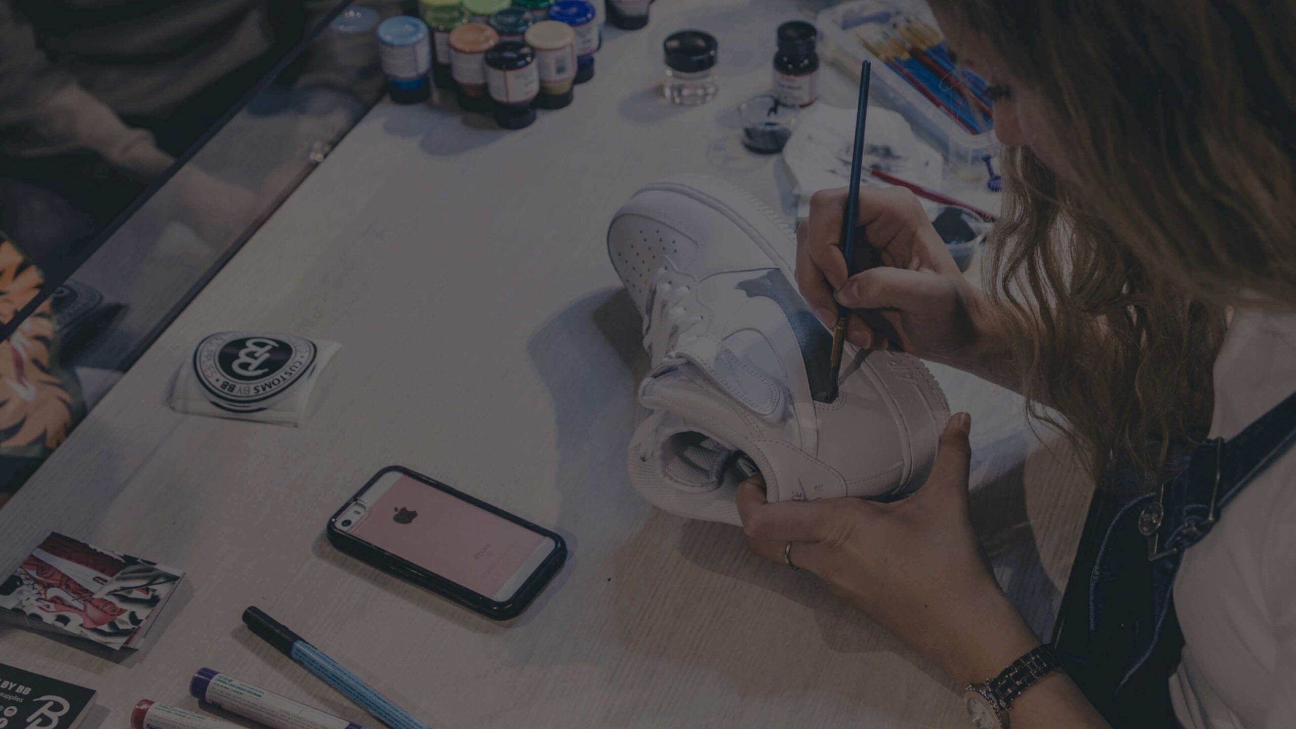 Tussen potten verf transformeert iemand een witte sneaker tot kunstwerk, geïnspireerd door Customs van BB op een nabijgelegen telefoon.