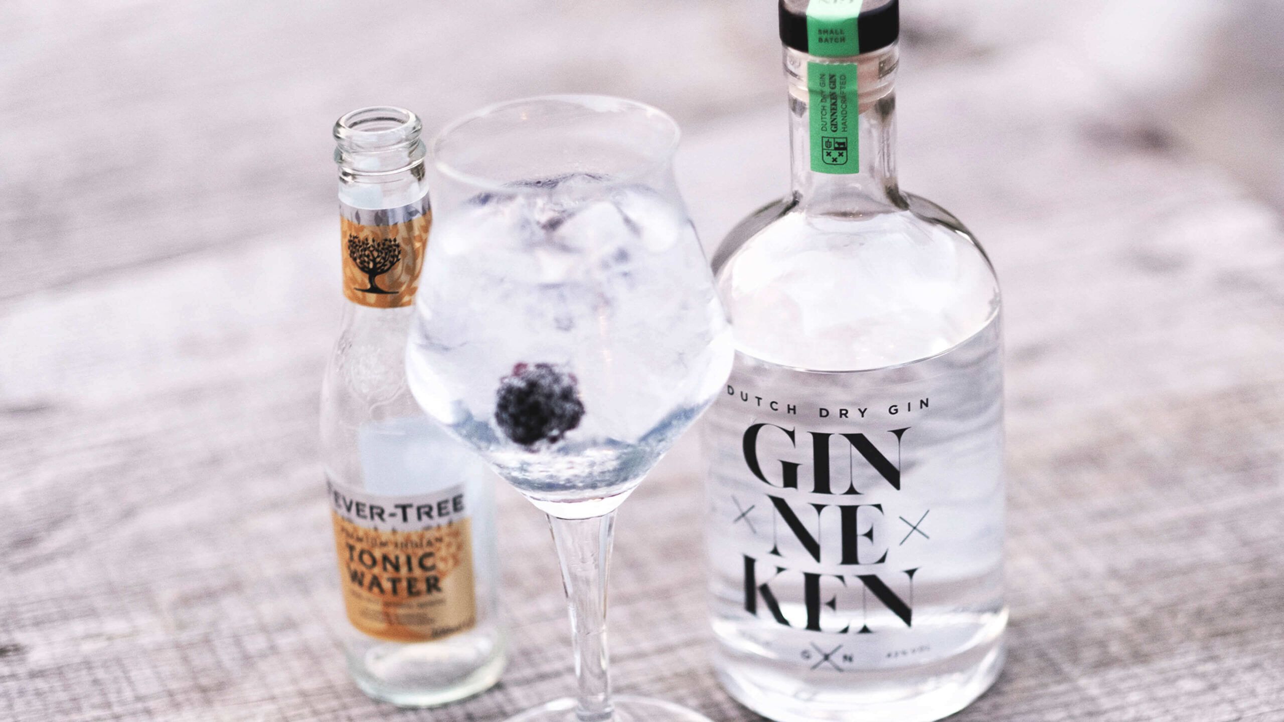 Close-up van een gin-tonic met ijs en bramen, op de achtergrond tonicflessen en GinnekenGin.com voor een eersteklas slokje.