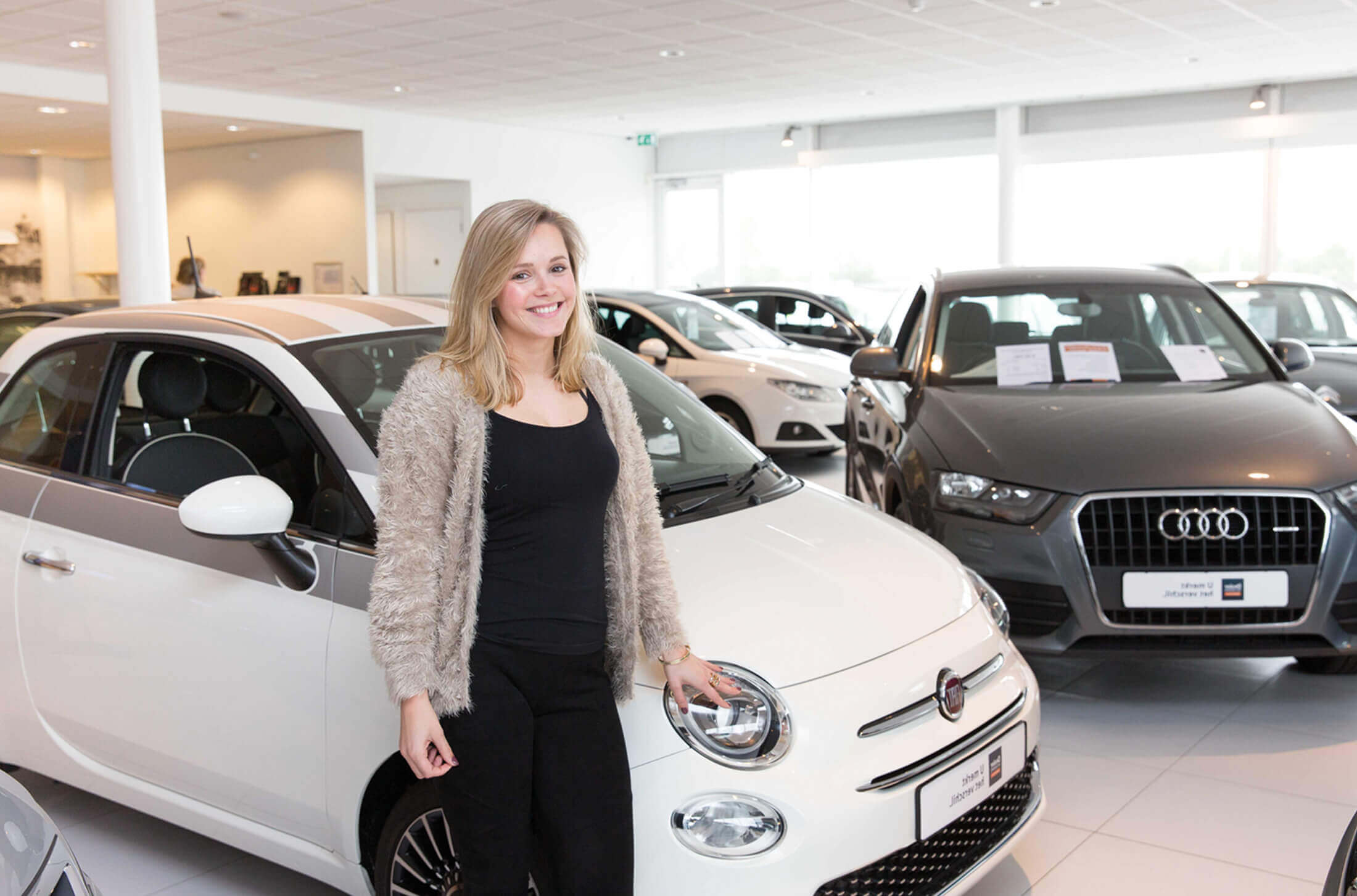 In een drukke showroom staat iemand naast een witte Fiat, met op de achtergrond gestroomlijnde auto's.
