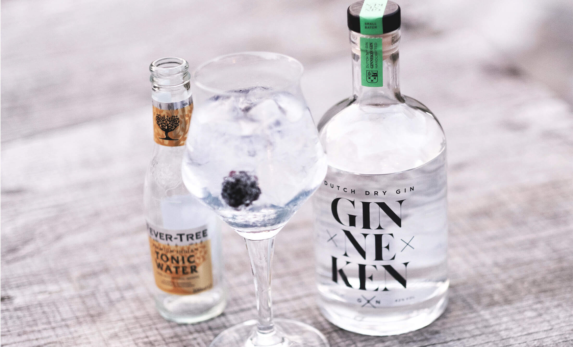 Een stijlvolle bramengin-tonic staat naast tonicwater en GinnekenGin op een rustiek houten oppervlak.