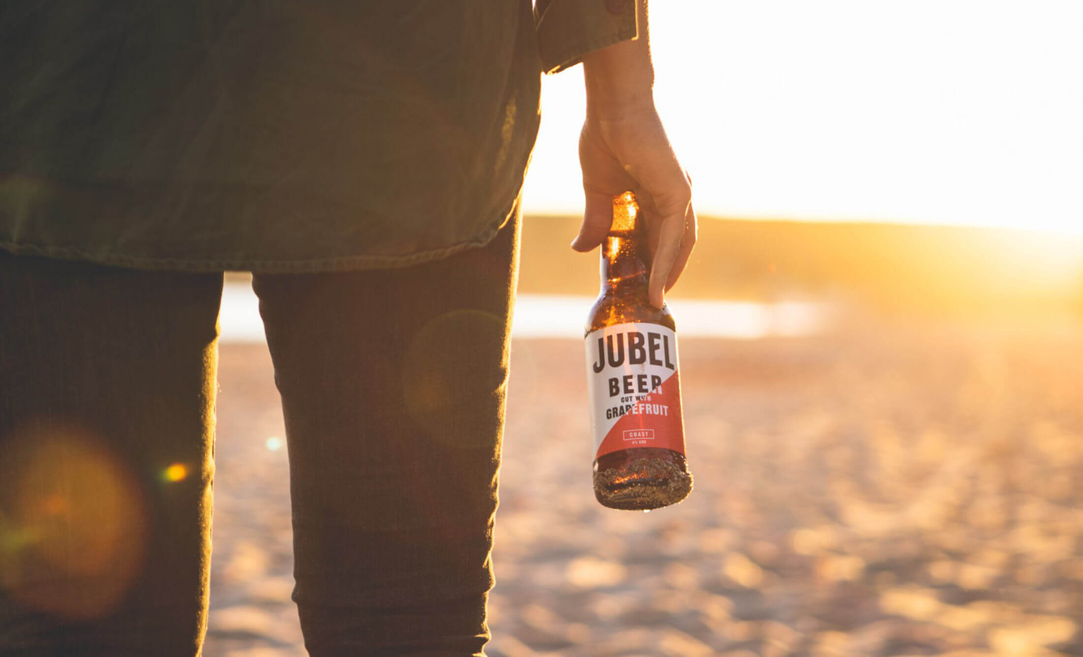 Terwijl de zon ondergaat en het strand goud kleurt, geniet iemand van een verfrissend Jubel-biertje.