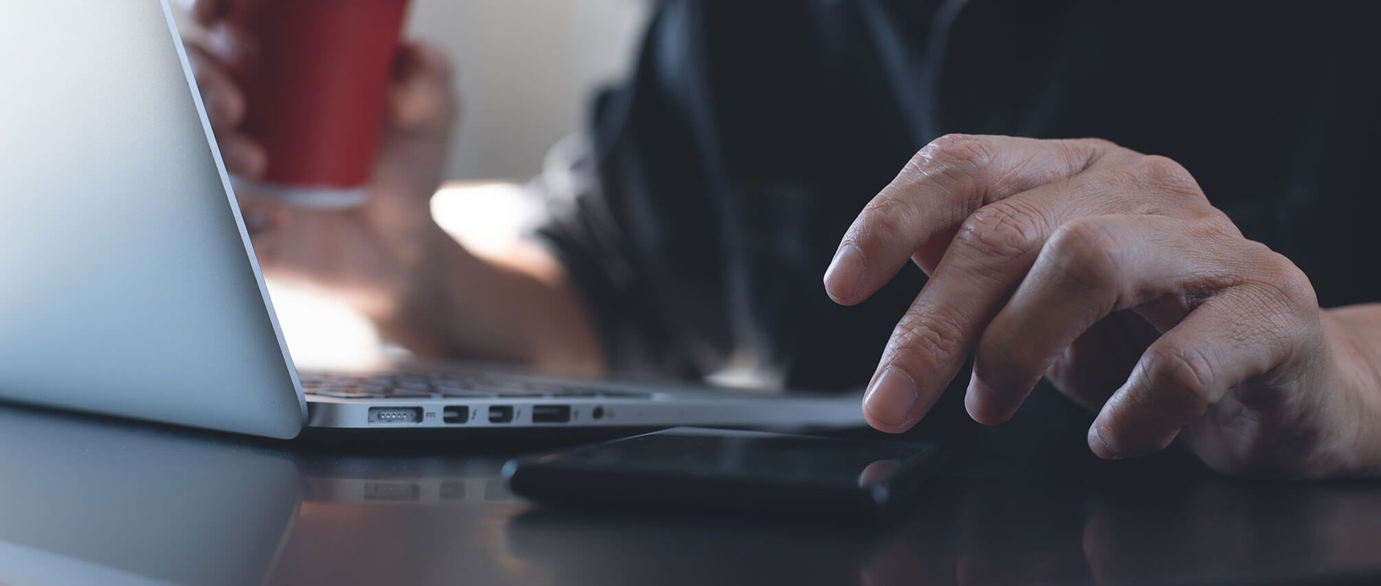 Laptop- en smartphonegebruiker aan een bureau, met een rode beker in zijn hand.