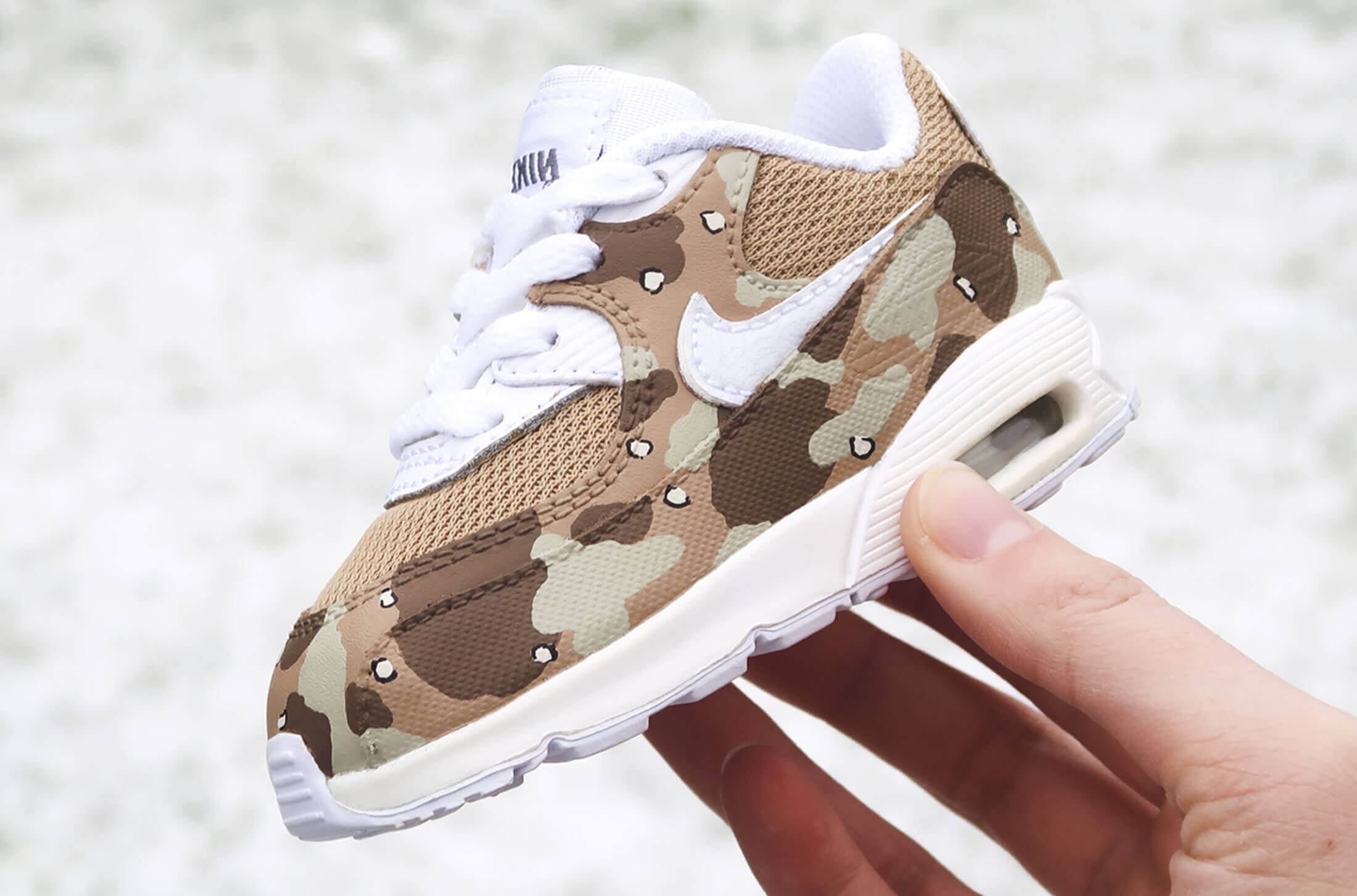 Een hand houdt een babysneaker met camouflageprint vast, met een witte swoosh en veters, speciaal gemaakt door BB.