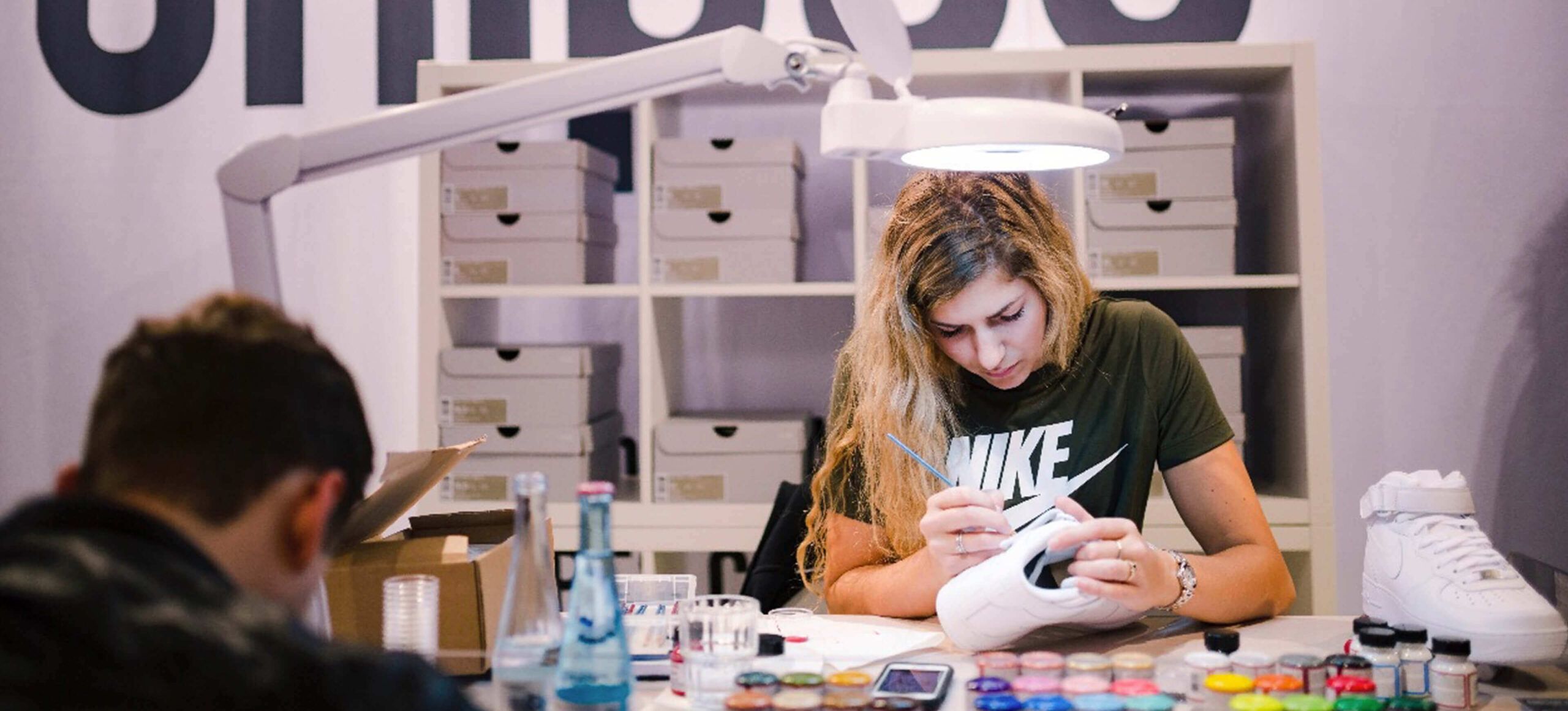 Een kunstenaar beschildert sneakers aan een felgekleurde tafel vol verf. Achterin zijn planken te zien die de chaos van "Customs by BB" laten zien.