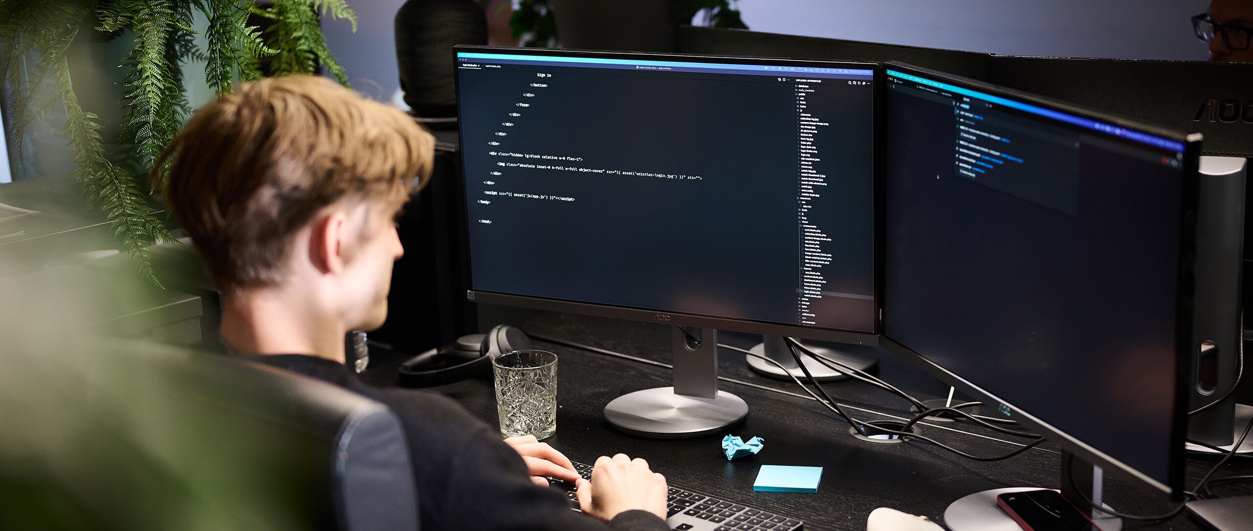 Een programmeur aan een bureau met twee monitoren, kantoorplanten en een glas water.