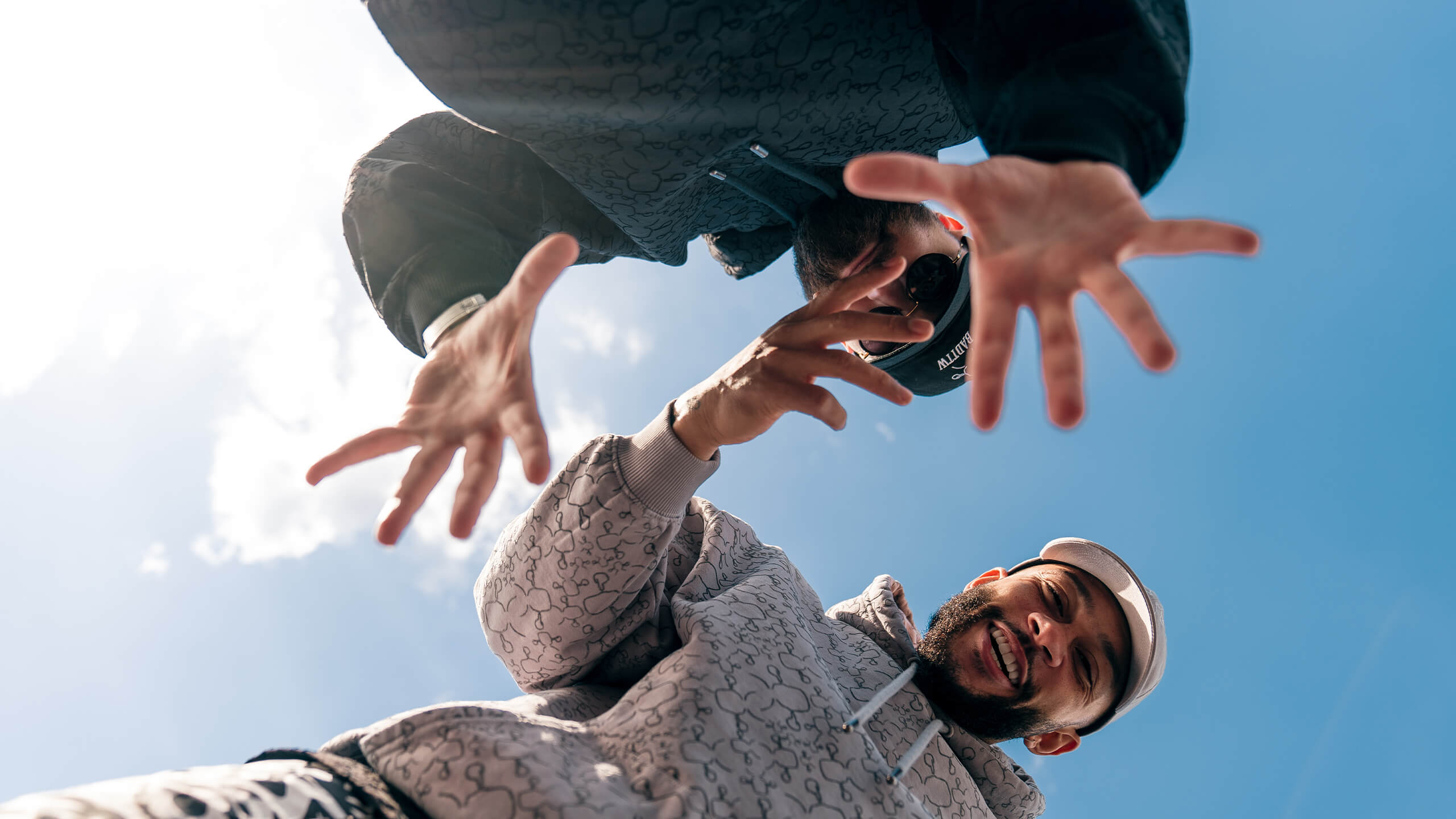 Twee figuren in hoodies strekken hun handen uit naar de camera, afstekend tegen een zonnige blauwe lucht.