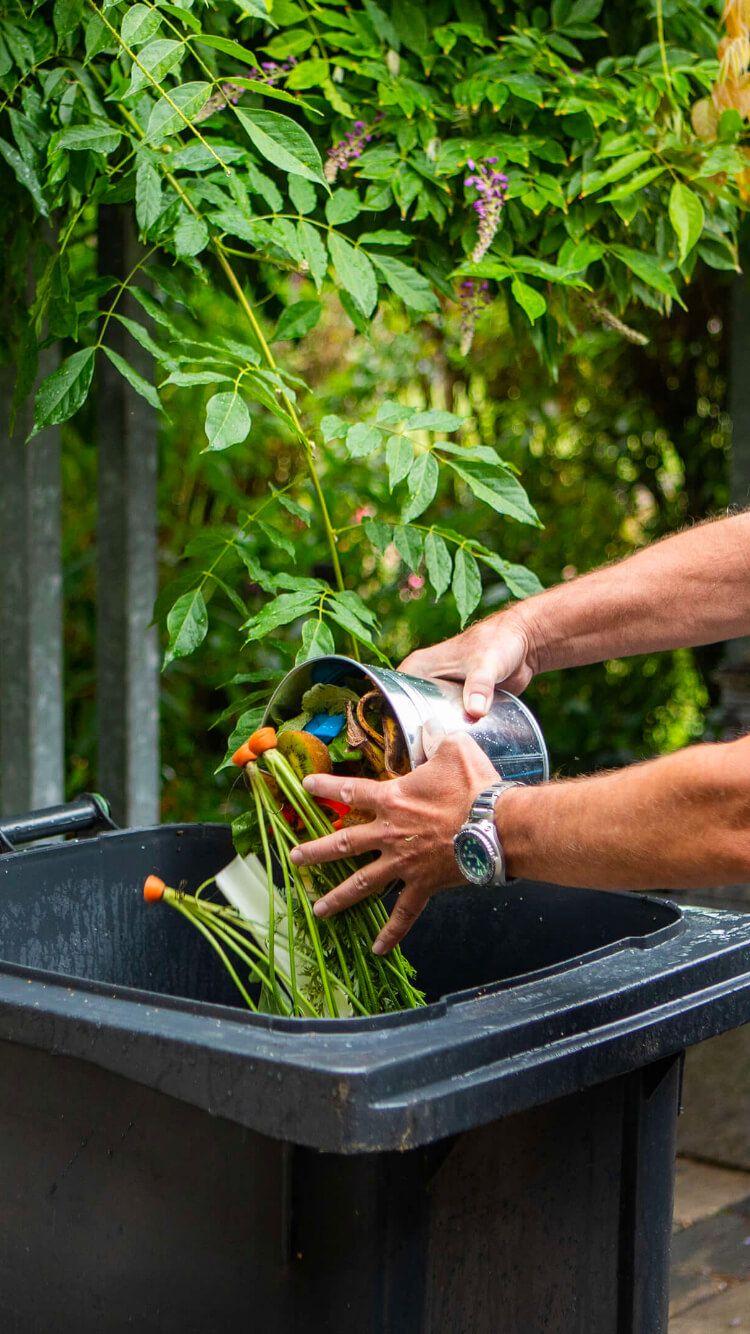 Enthousiast kieperen ze het plantenafval van een metalen bak naar een grote zwarte composthoop voor Samen Aan De Bak!.