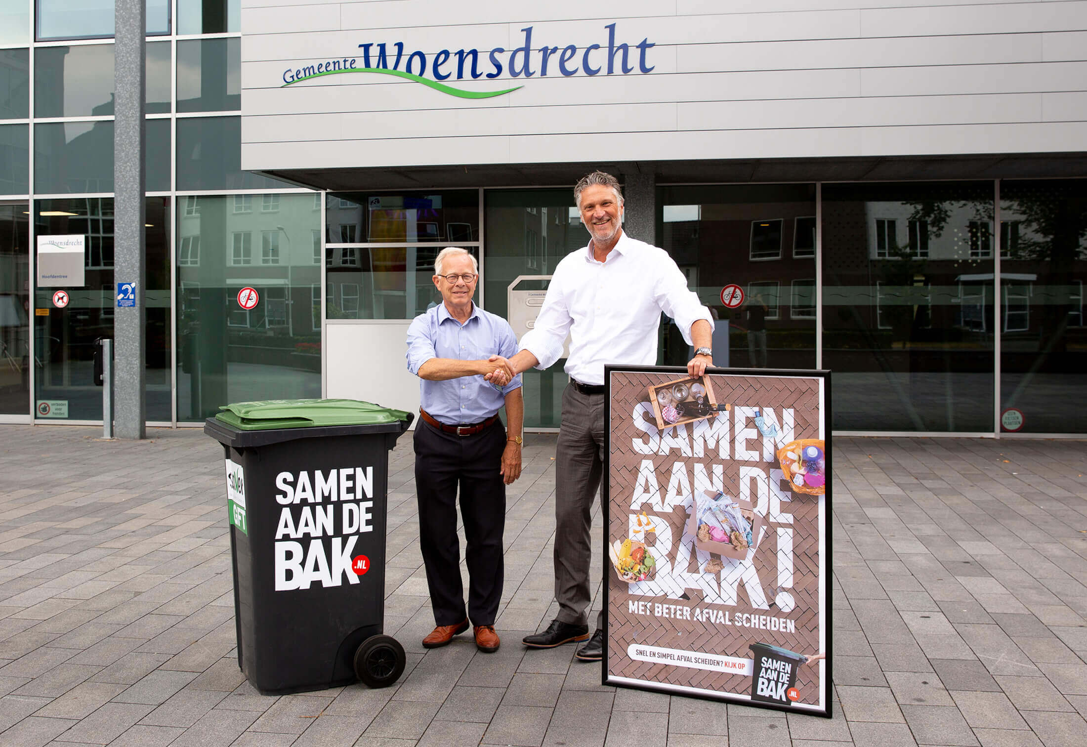 Twee mannen schudden elkaar de hand bij een prullenbak en "Samen Aan De Bak!" affiche in Woensdrecht.