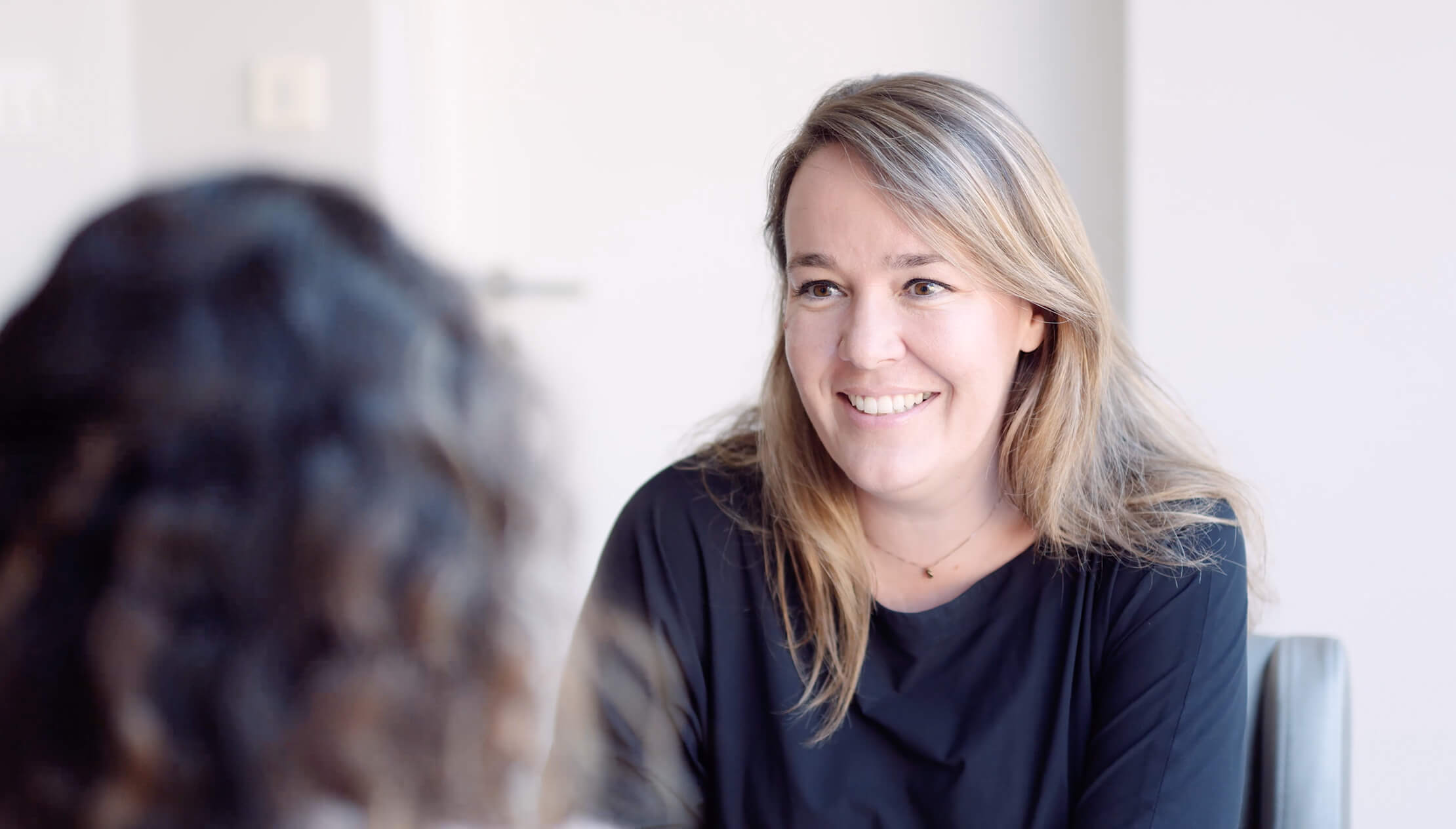 Een langharige vrouw lacht hartelijk naar iemand en legt daarmee de gastvrije sfeer van CS Wonen vast.