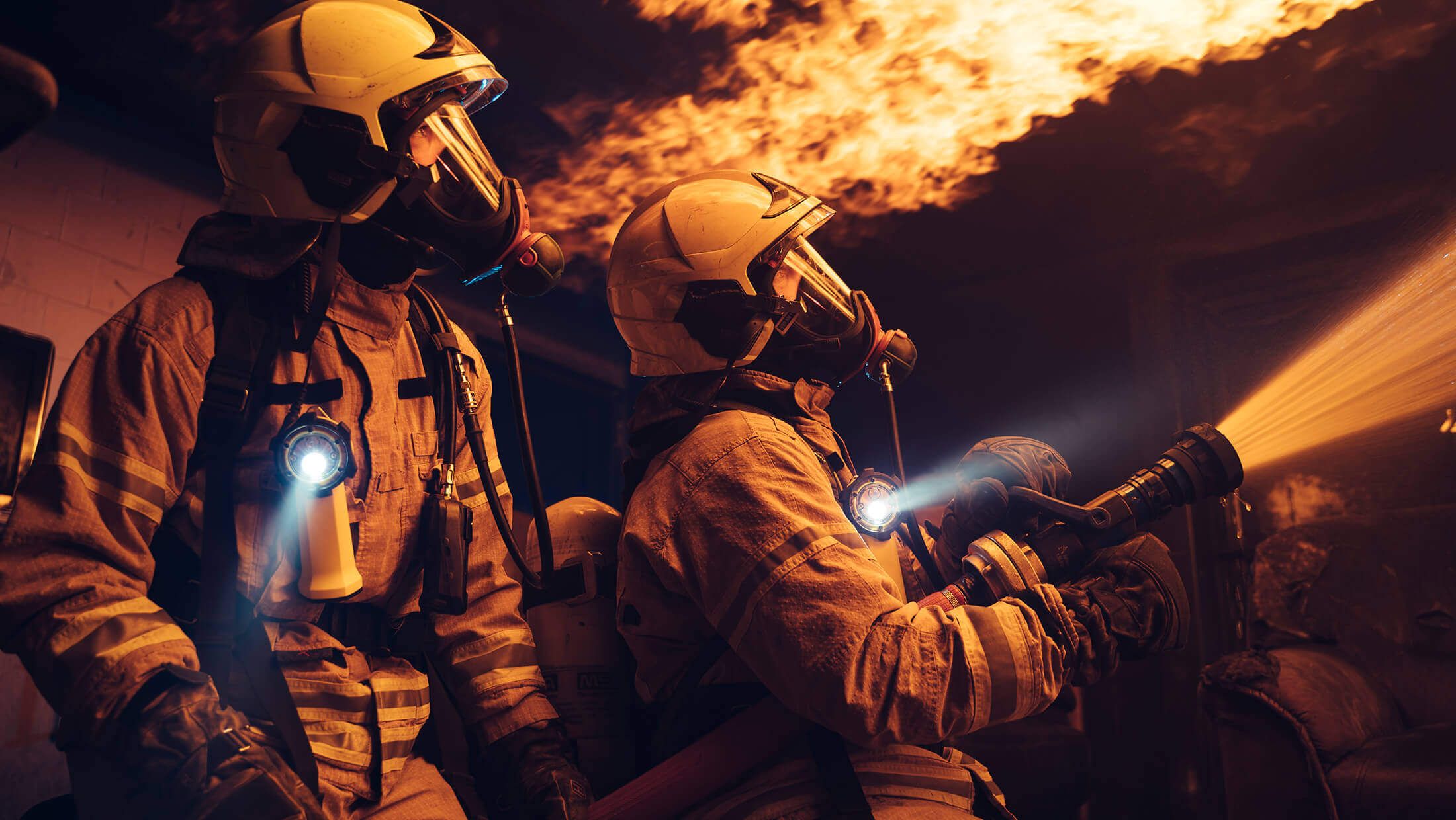 Brandweerlieden in uniform blussen vakkundig een brand in een gebouw, aan de hand van flakkerende vlammen en de stralen van zaklampen.