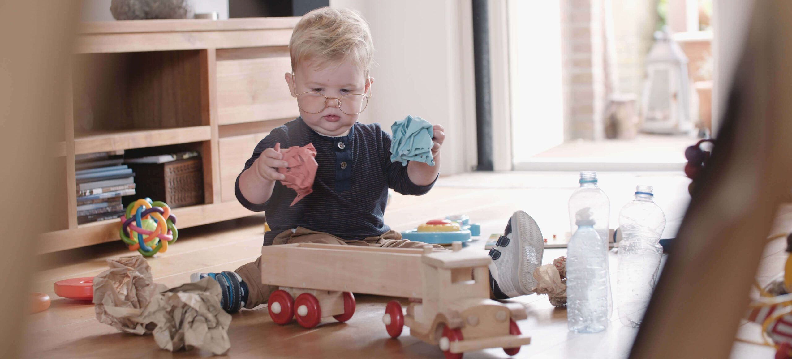 Een peuter verkent vrolijk een kamer vol speelgoed met een houten vrachtwagen en papier, wat helemaal in de sfeer van "Samen Aan De Bak!" komt.