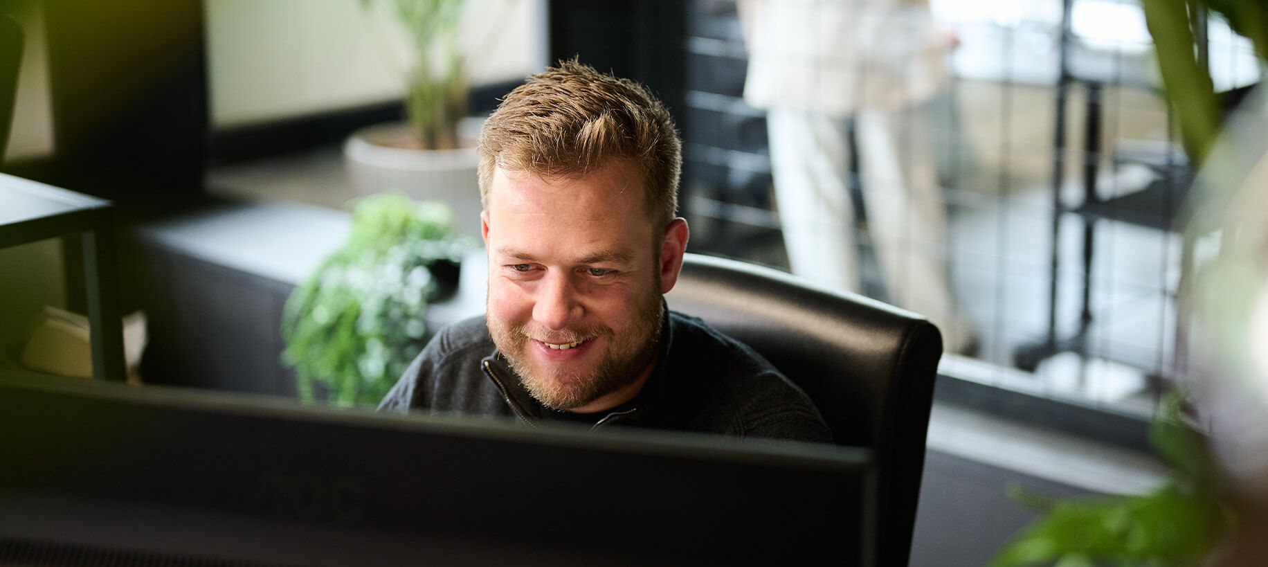 Een man grijnst naar zijn computer in een kantoor vol planten. Hij straalt succes uit.