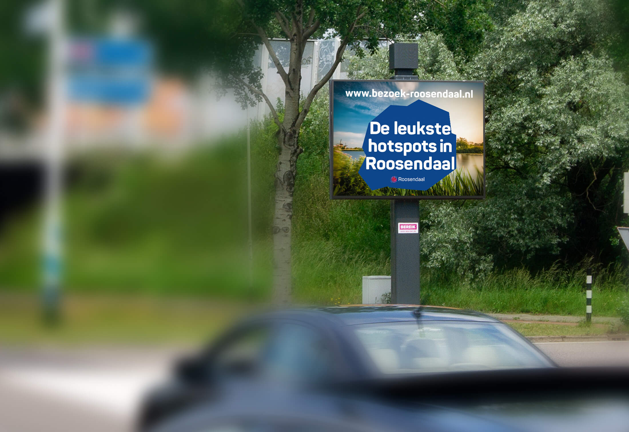 Reclamebord toont "Toplocaties in Roosendaal" met een link naar de website; onscherpe auto op de voorgrond.