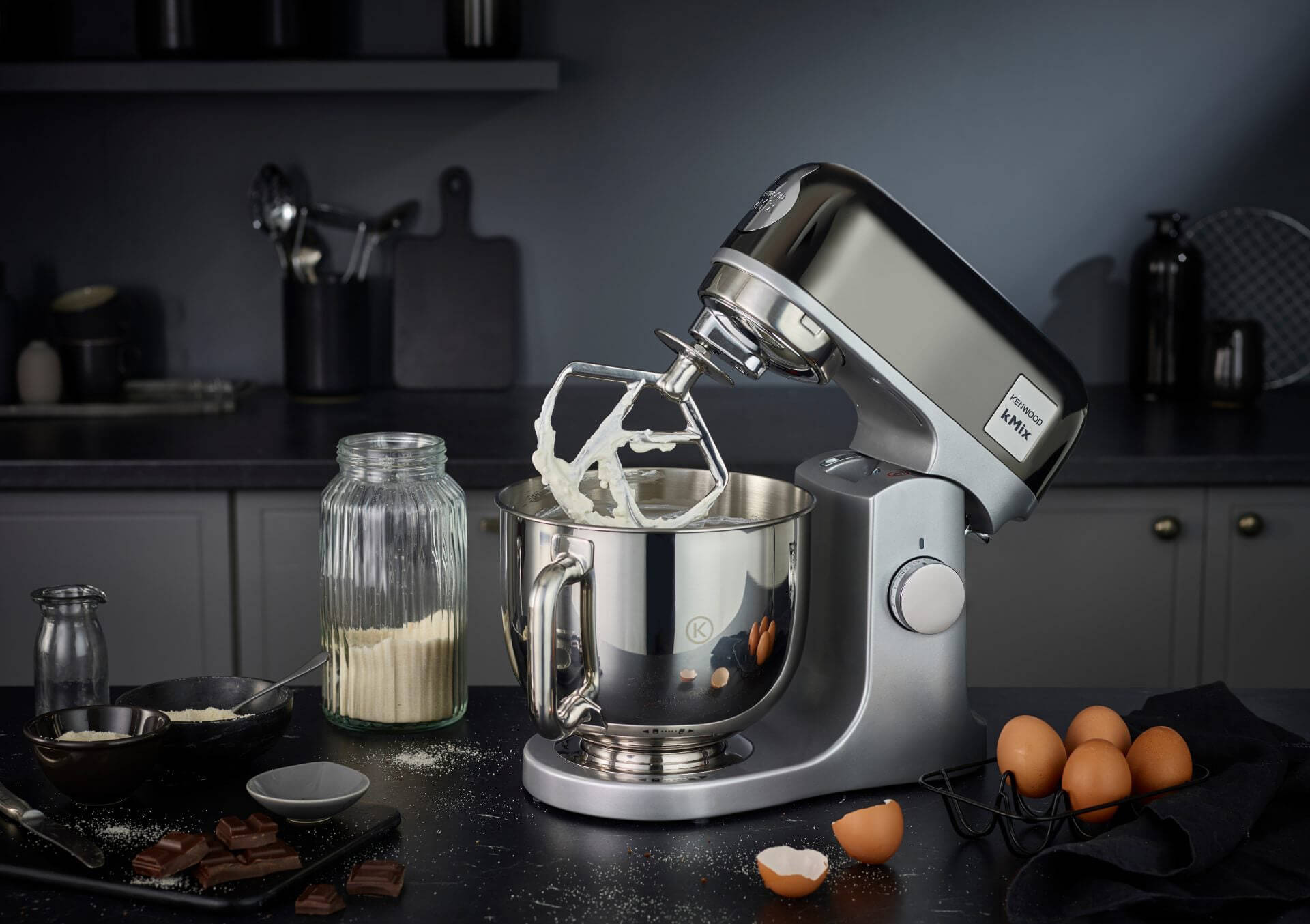 Zilveren mixer met slagroom in een keuken, omringd door eieren, bloem en chocolade.