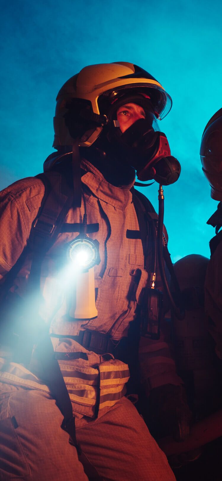 Een brandweerman in volledige uitrusting met een zaklamp staat zelfverzekerd tegen een rokerige, blauw verlichte achtergrond.