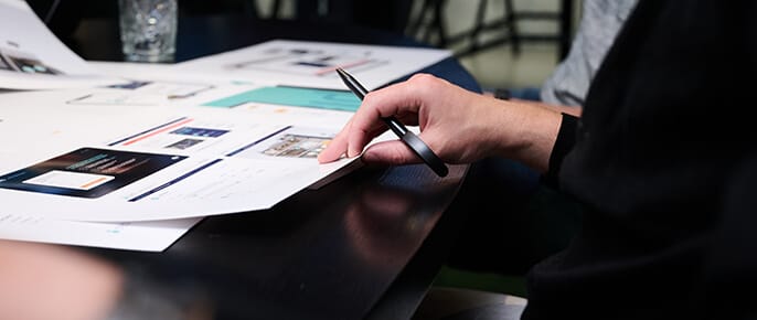Iemand zit aan een tafel, met pen in de hand, huisstijldocumentatie en ontwerptekeningen te bekijken.
