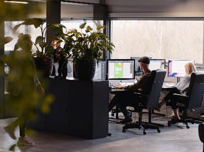 Modern kantoor: mensen achter bureaus, actieve computers, grote potplanten op de voorgrond.