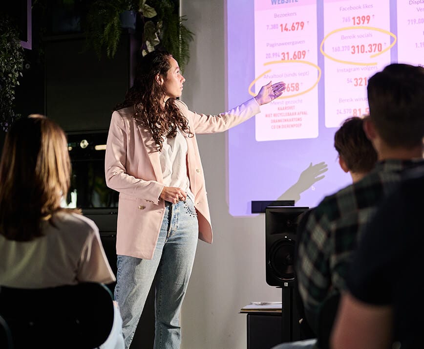 Een vrouw in een blazer en spijkerbroek presenteert gegevens en wijst naar getallen op een geprojecteerde dia.