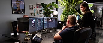 Twee ontwikkelaars werken aan computerbureaus met meerdere beeldschermen, groene planten en een chique kantoorinrichting.