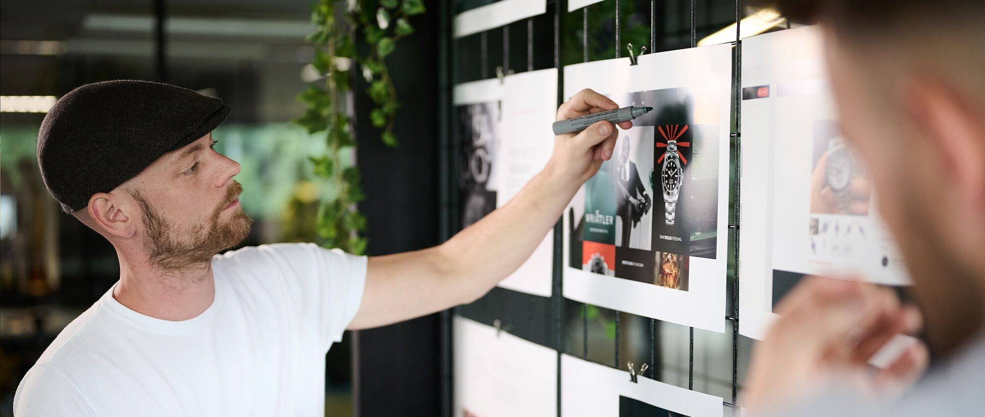 Een man met een pet schetst druk ideeën op een ontwerp dat op de rastermuur is geprikt.