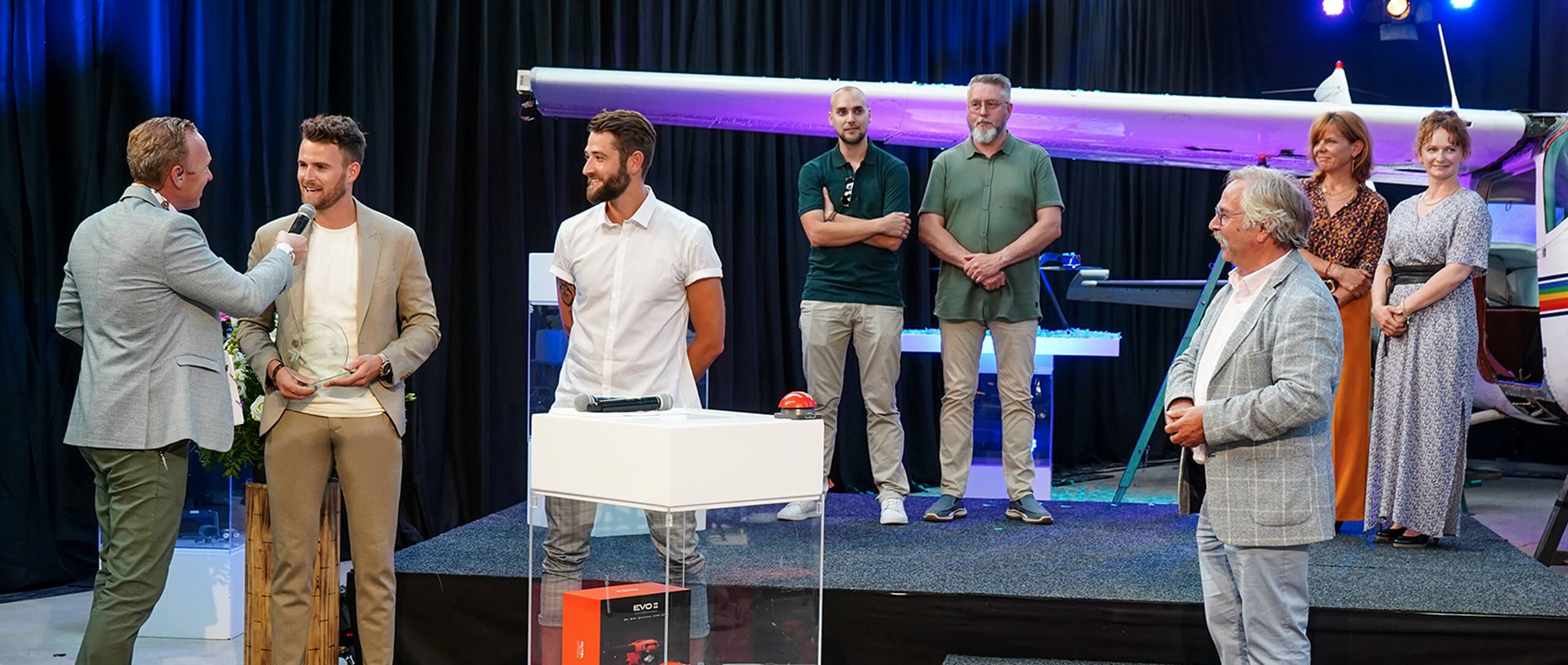 Op het podium praten twee mannen met een microfoon, voor een klein vliegtuig en een groep.