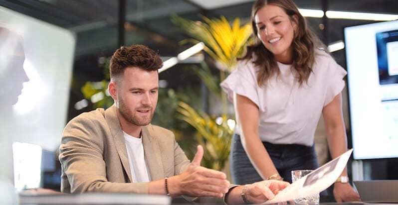 Een man en een vrouw bespreken marketingcampagnes aan een strakke tafel, omringd door weelderige planten in een modern kantoor.