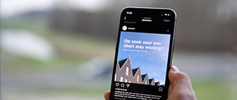 Op een smartphone zie je huizen met puntgevels onder een blauwe lucht, terwijl Nederlandse teksten hun charme benadrukken.