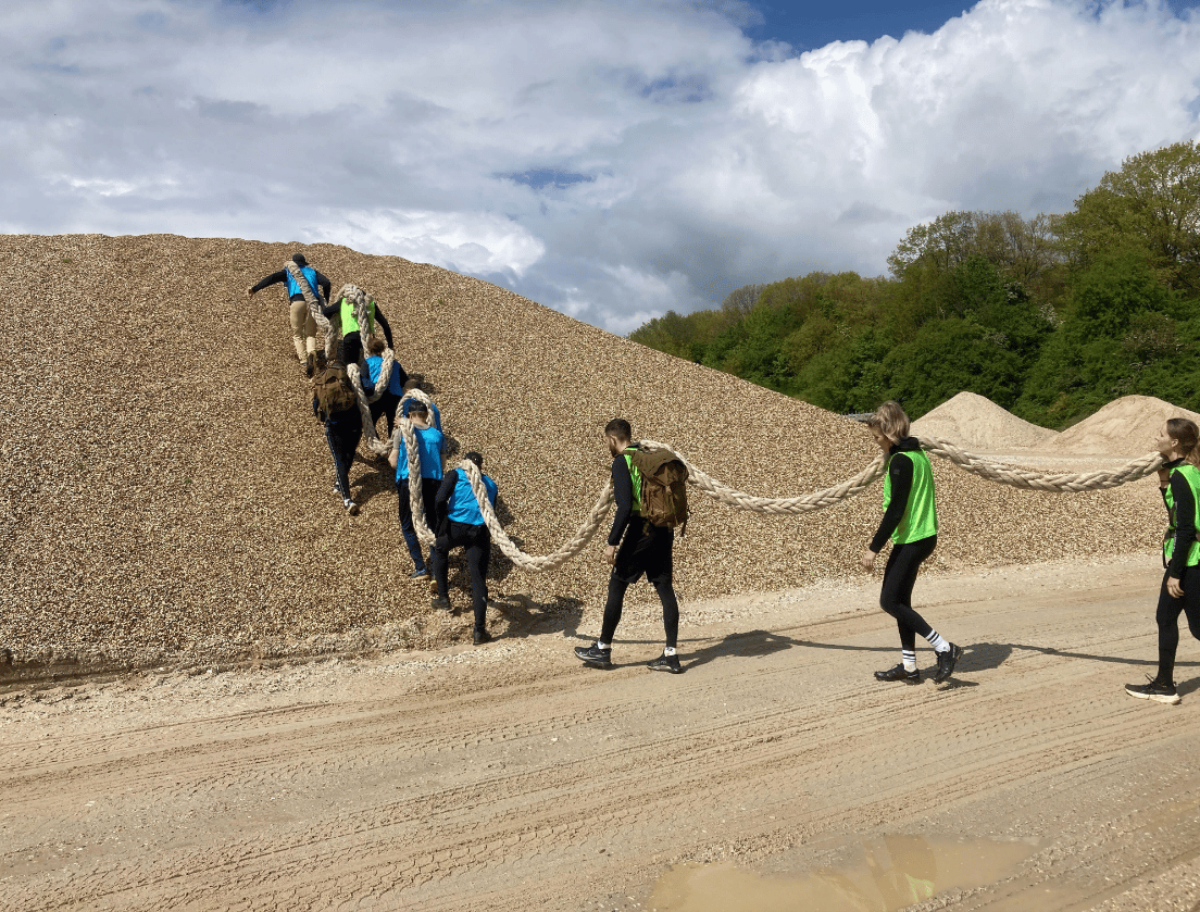 Een groep klimt in één rij een grindheuvel op, allemaal vasthoudend aan hetzelfde dikke touw.