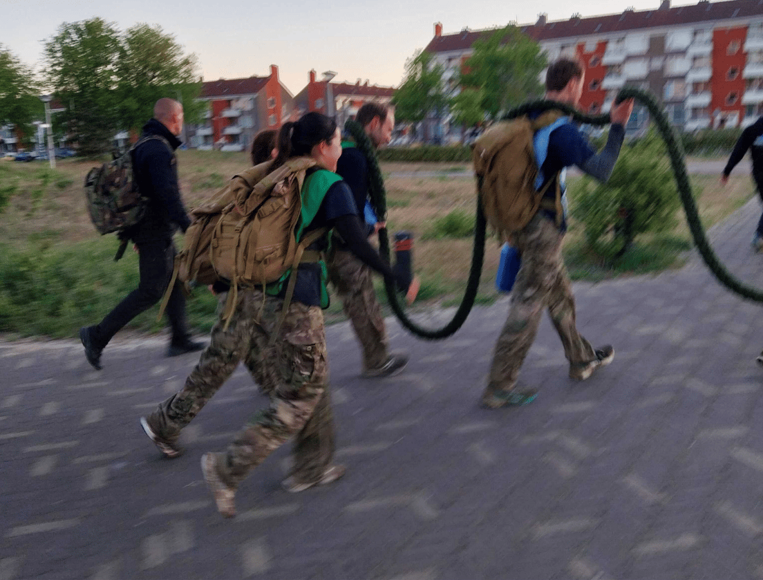 Vijf mensen in camouflagebroeken en rugzakken wandelen samen, terwijl ze een lang, zwaar touw dragen.