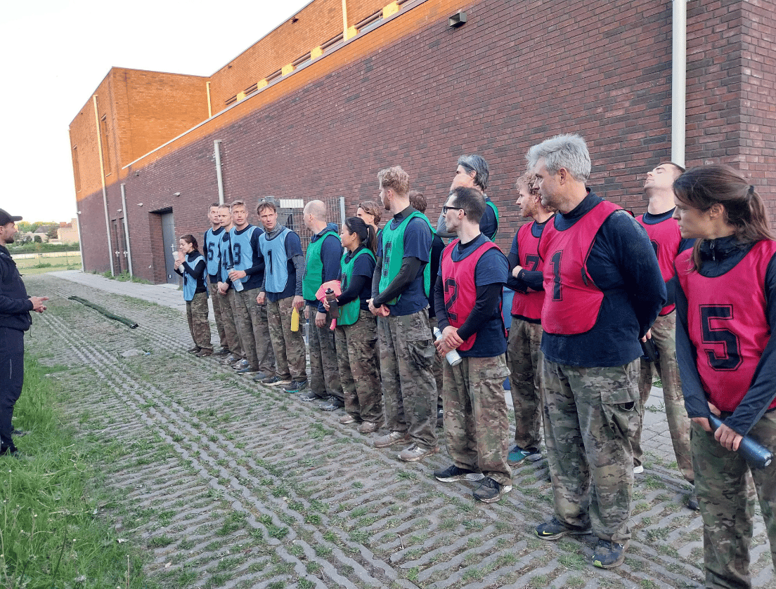 Een groep in camouflagebroeken en vesten luistert naar iemand in het zwart die buiten instructies geeft.