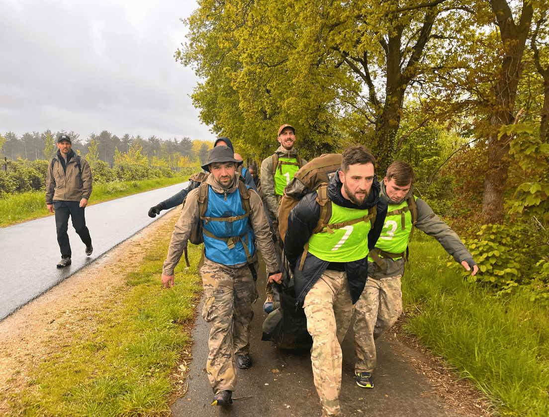 Vier mannen in camouflagekleding trekken een zware last over een plattelandspad; een ander volgt in de regen.