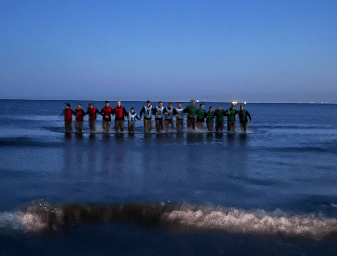 Mensen in felgekleurde shirts staan in ondiep zeewater bij zonsondergang, onder een blauwe lucht.