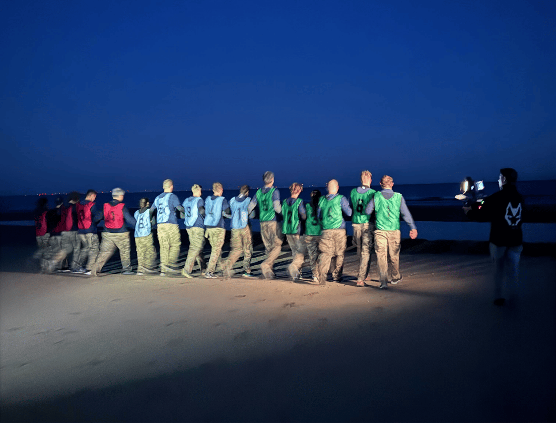 Mensen in kleurrijke shirts lopen 's avonds op het strand in één rij, waarbij één persoon de weg wijst.