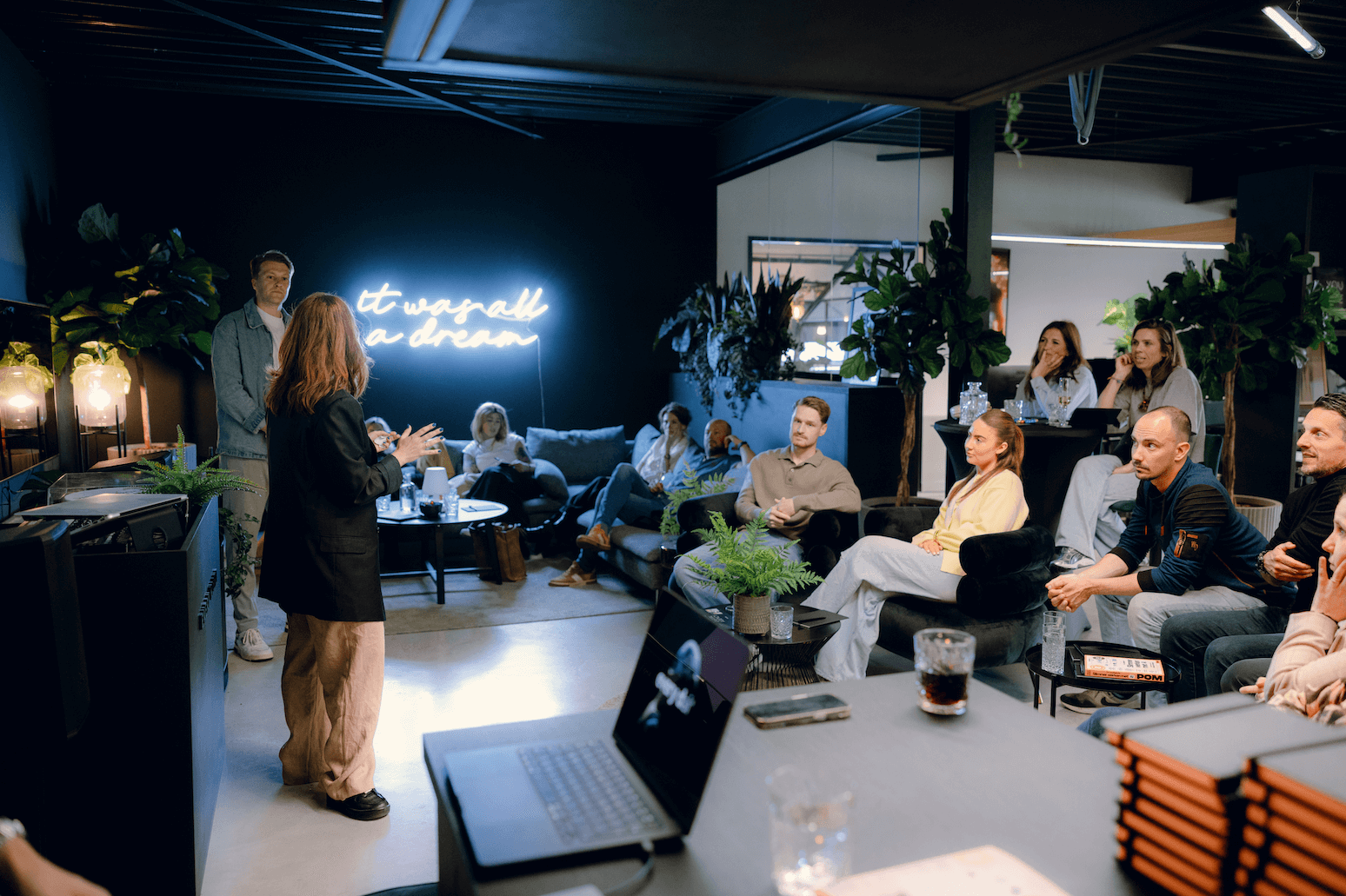 Een vrouw presenteert aan een zittende groep in een modern kantoor met een neonbord: "het was allemaal een droom.
