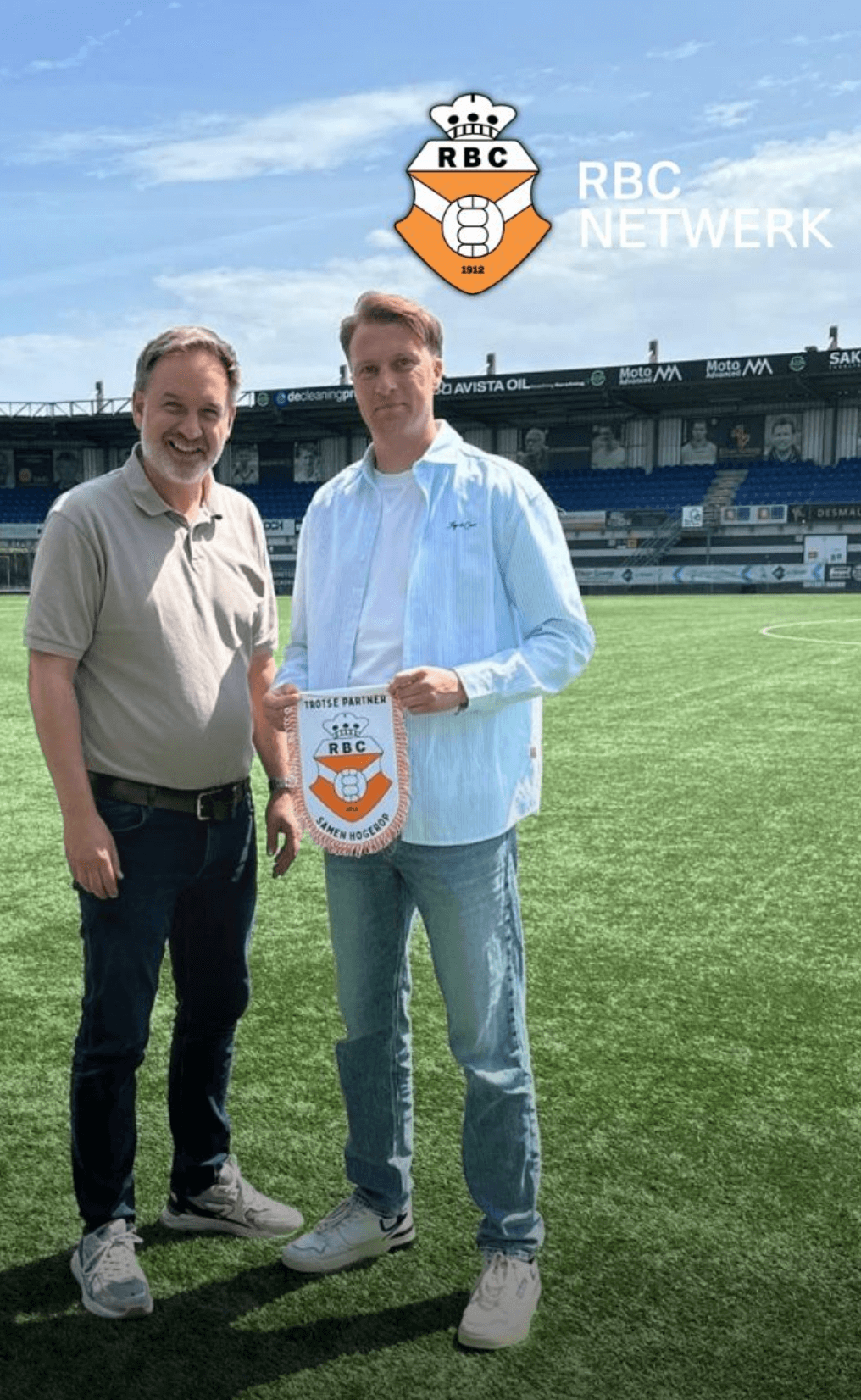 Twee mannen op een voetbalveld houden een RBC Netwerk bord vast, stadion en logo zichtbaar op de achtergrond.