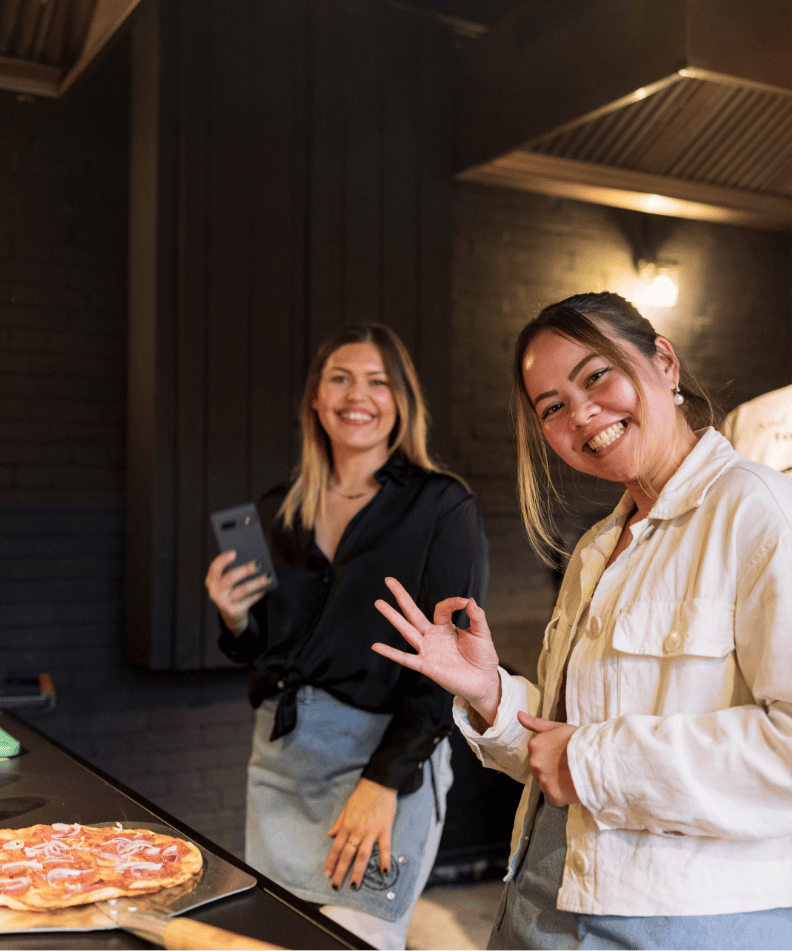 Twee mensen in schorten lachen terwijl ze pizza maken; de een maakt een foto, de ander geeft een OK-teken. 🍕📸👌.