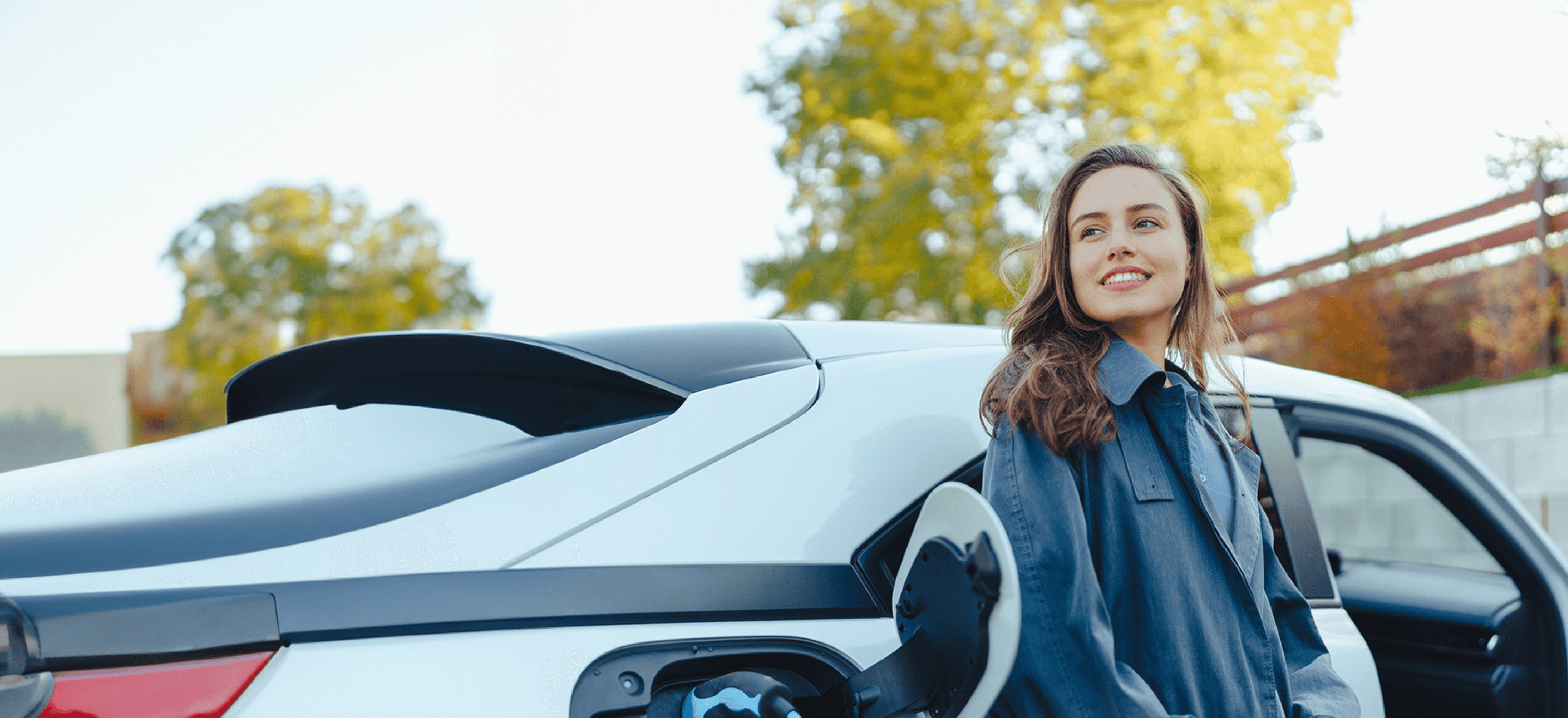 Een vrouw glimlacht naast een opladende witte elektrische auto, met bomen op de achtergrond.