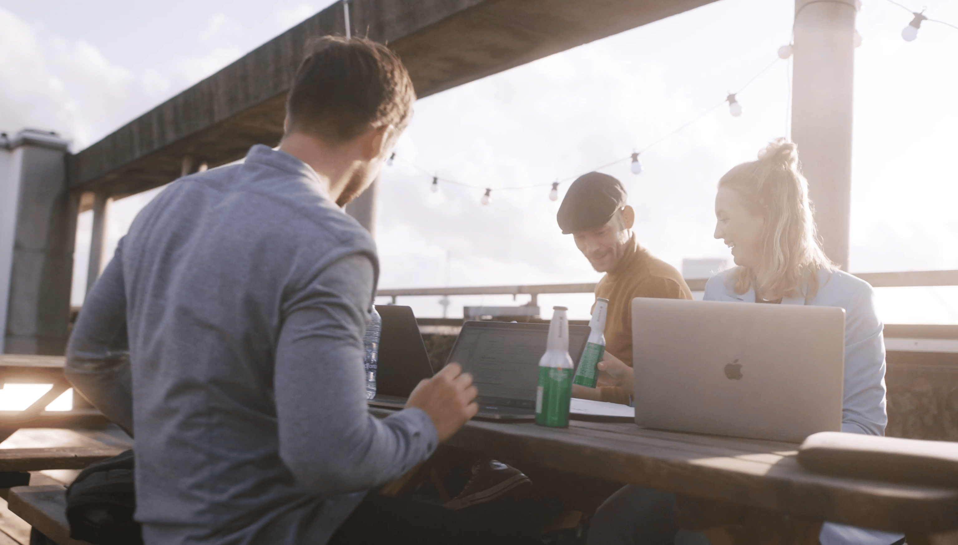 Drie mensen kletsen en lachen rond hun laptops aan een tafeltje buiten, met daglicht en lichtslingers erboven.