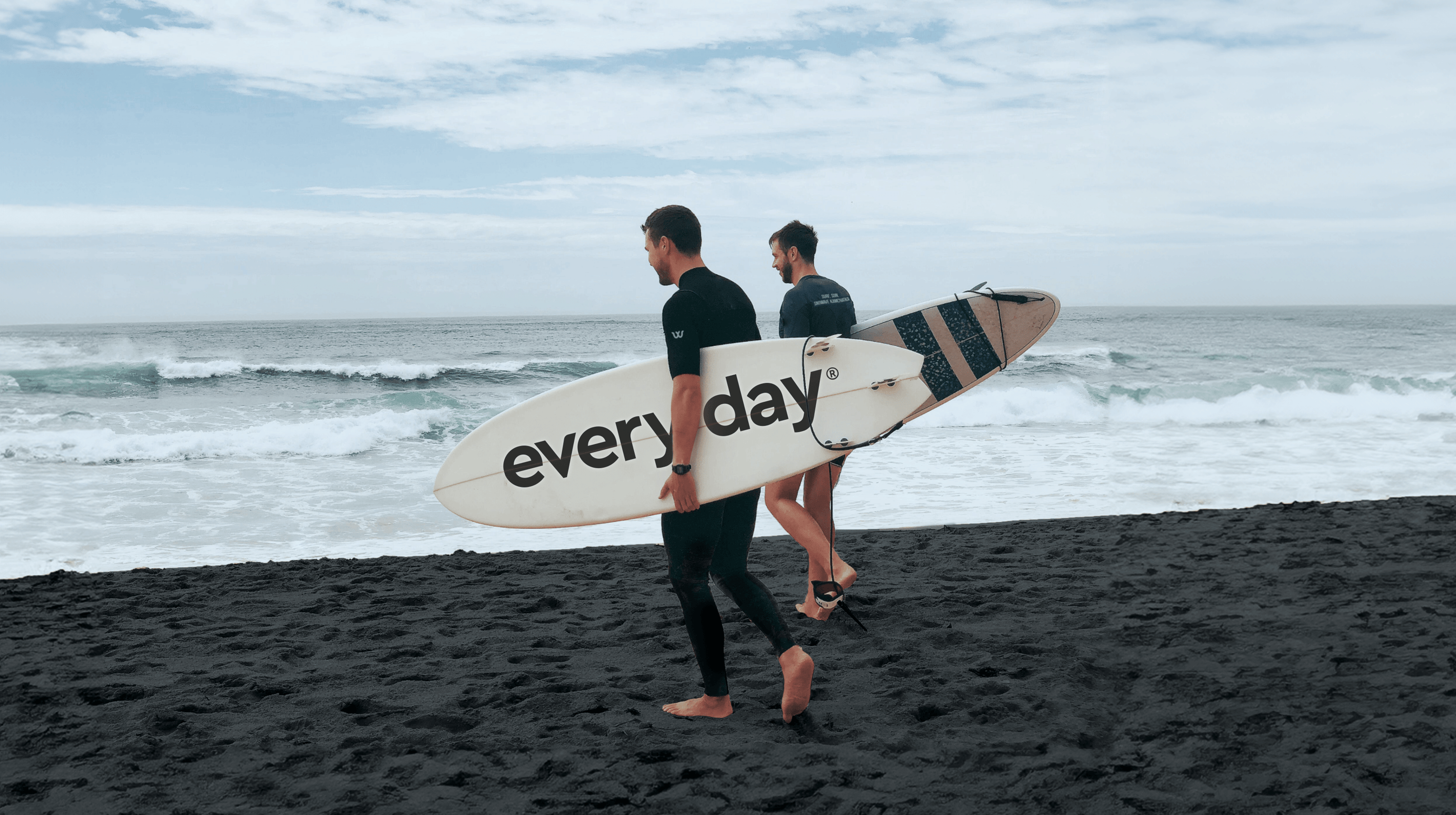 Twee surfers wandelen over een zwart zandstrand, met een surfplank in hun handen met het opschrift 'elke dag'.