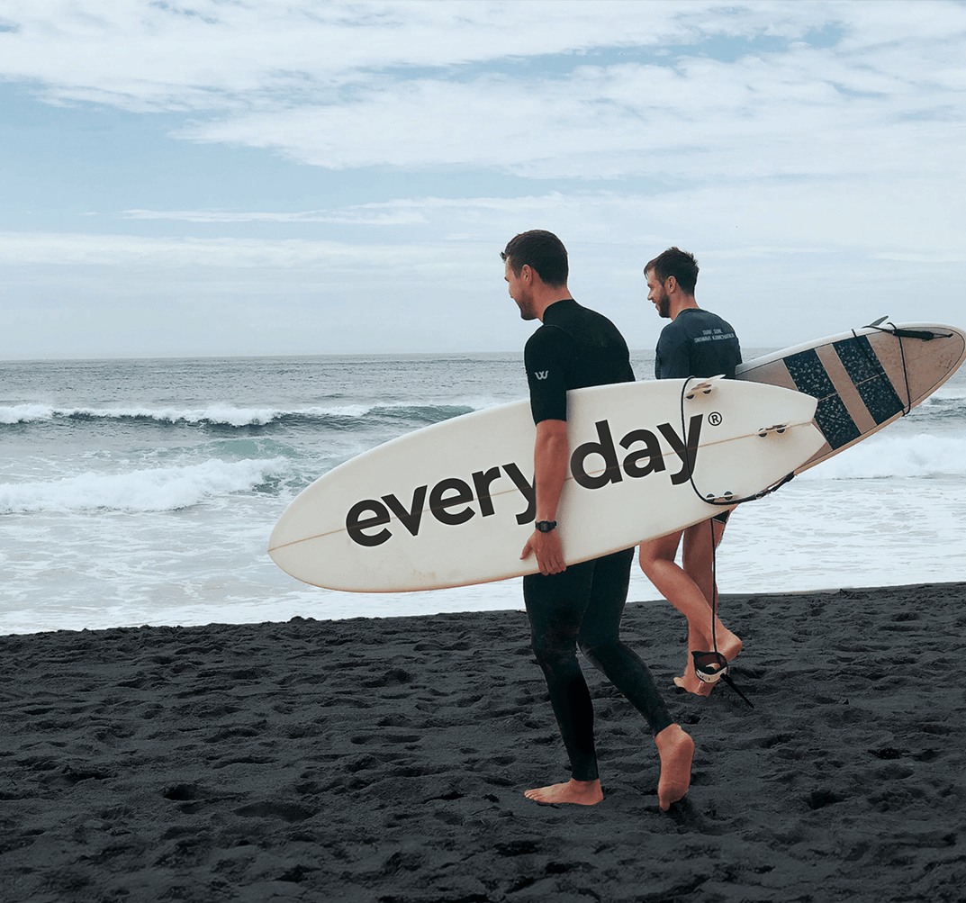 Twee surfers gaan naar de oceaan. De één houdt een bord vast met het opschrift 'elke dag'.